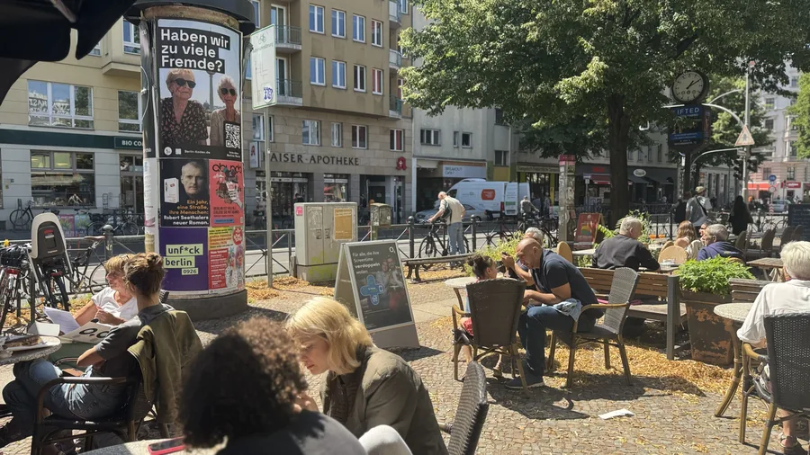 Straßencafé mit einer Litfaßsäule und dem Berlin-Kodex-Motiv Haben wir zu viele Fremde?